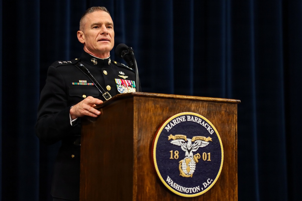 A U.S. Marine Corps officer in dress blues uniform is speaking at a podium at Marine Barracks Washington, D.C. The officer displays multiple medals and ribbons and appears to hold a general officer rank based on visible star insignia on the shoulders. The setting appears to be a formal military ceremony or official event.