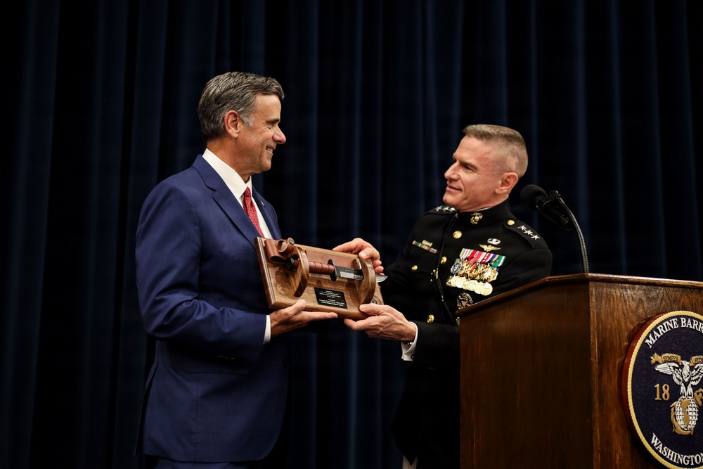 A U.S. Marine Corps officer in dress uniform is presenting a wooden plaque/award to a civilian man in a blue suit at what appears to be a formal ceremony at Marine Barracks Washington. The military officer displays two stars on his collar indicating the rank of Major General, and wears numerous service ribbons and medals. The event is taking place at a podium bearing the Marine Barracks Washington seal.