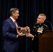 A U.S. Marine Corps officer in dress uniform is presenting a wooden plaque/award to a civilian man in a blue suit at what appears to be a formal ceremony at Marine Barracks Washington. The military officer displays two stars on his collar indicating the rank of Major General, and wears numerous service ribbons and medals. The event is taking place at a podium bearing the Marine Barracks Washington seal.