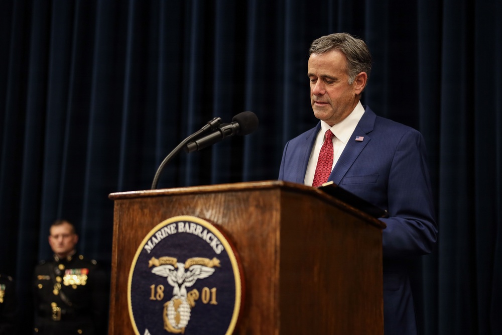 A man in a blue suit with a red tie and American flag lapel pin stands at a wooden podium bearing the 'Marine Barracks 1801' seal, appearing to read from notes or a document. A uniformed U.S. Marine officer with medals and ribbons is visible in the background. The setting appears to be a formal military ceremony or official event.