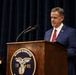 A man in a blue suit with a red tie and American flag lapel pin stands at a wooden podium bearing the 'Marine Barracks 1801' seal, appearing to read from notes or a document. A uniformed U.S. Marine officer with medals and ribbons is visible in the background. The setting appears to be a formal military ceremony or official event.