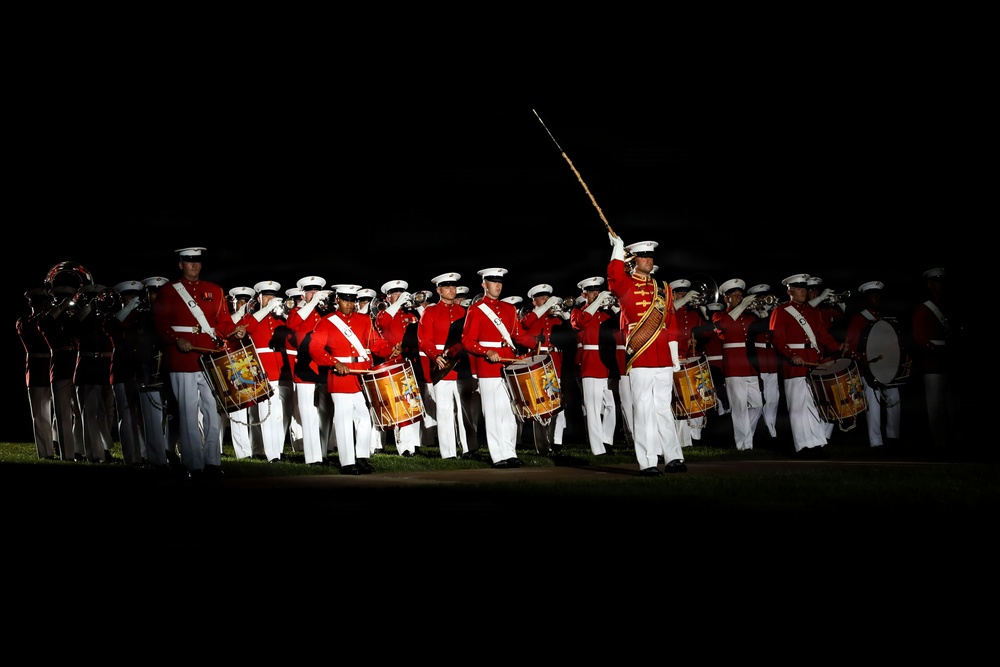 A military marching band performs an evening ceremony, illuminated against a dark background. The musicians are dressed in distinctive red tunics with white trousers and white peaked caps, consistent with U.S. Marine Corps dress uniform. A drum major leads the formation with a raised mace, while drummers and brass players are visible in formation.