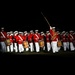 A military marching band performs an evening ceremony, illuminated against a dark background. The musicians are dressed in distinctive red tunics with white trousers and white peaked caps, consistent with U.S. Marine Corps dress uniform. A drum major leads the formation with a raised mace, while drummers and brass players are visible in formation.
