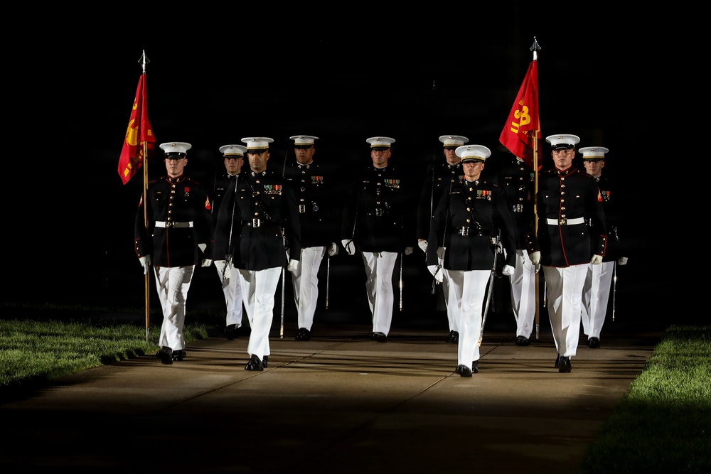 A formation of United States Marines in dress blue uniforms marches in a ceremonial parade at night, illuminated by artificial lighting against a dark background. Two Marines carry red guidon flags with gold Marine Corps insignia on either flank of the formation. The precision marching and formal attire suggest this is an official military ceremony, likely an Evening Parade or similar formal event.