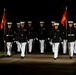 A formation of United States Marines in dress blue uniforms marches in a ceremonial parade at night, illuminated by artificial lighting against a dark background. Two Marines carry red guidon flags with gold Marine Corps insignia on either flank of the formation. The precision marching and formal attire suggest this is an official military ceremony, likely an Evening Parade or similar formal event.