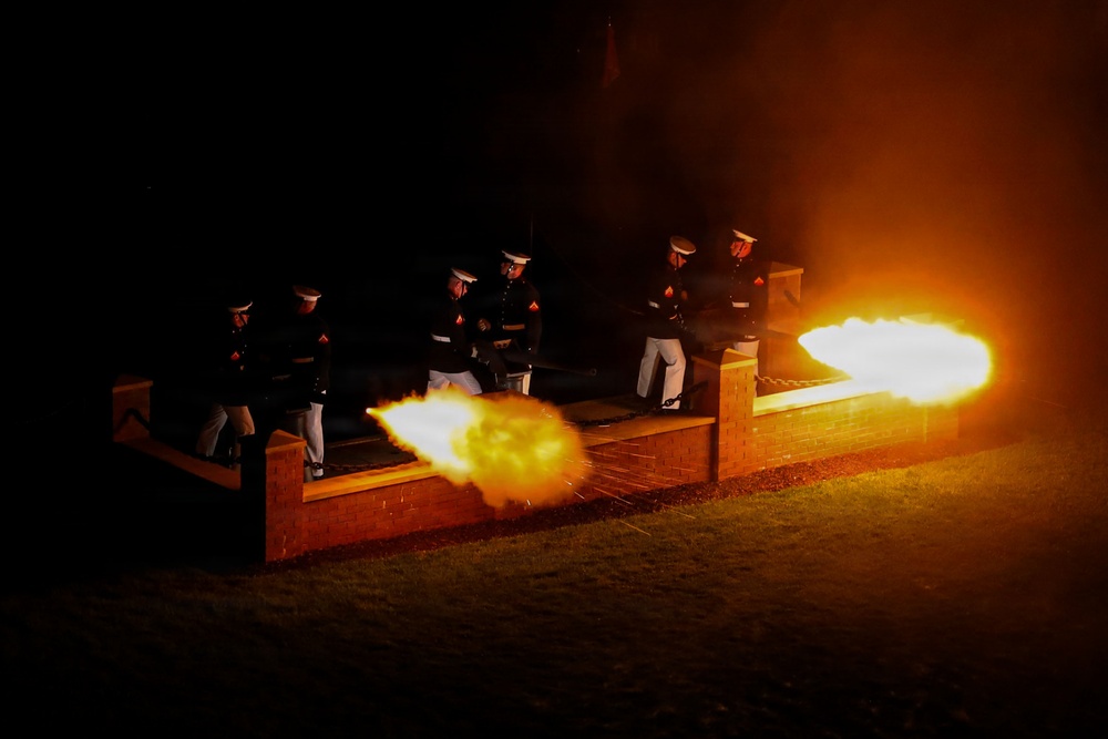 A nighttime military ceremonial cannon firing is taking place, with uniformed personnel standing behind a low brick wall structure. Two cannons are visibly firing simultaneously, producing large orange and white muzzle blasts. The scene is dramatically illuminated solely by the cannon fire against a completely dark background.
