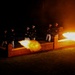 A nighttime military ceremonial cannon firing is taking place, with uniformed personnel standing behind a low brick wall structure. Two cannons are visibly firing simultaneously, producing large orange and white muzzle blasts. The scene is dramatically illuminated solely by the cannon fire against a completely dark background.
