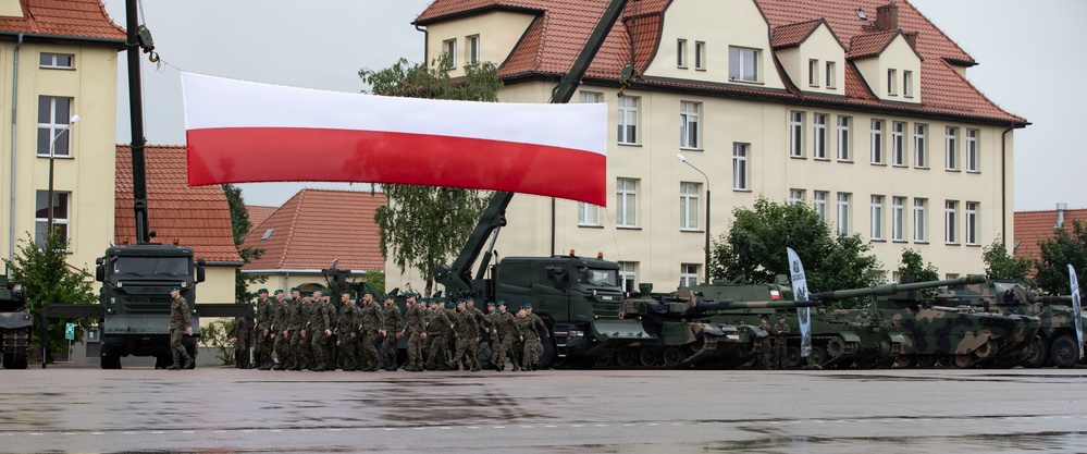 A military parade or ceremony is taking place on a wet parade ground, with soldiers marching in formation beneath a large Polish national flag (white and red horizontal stripes) suspended by a crane. Several military vehicles, including what appear to be Leopard 2 tanks and military trucks, are displayed in the background. The setting appears to be a military base or garrison, with institutional buildings visible in the background.