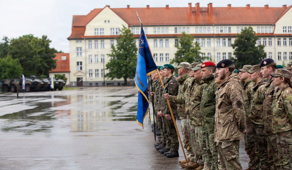 A multinational military formation is standing at attention on a wet parade ground, holding a blue NATO flag with gold fringe. Soldiers wearing different camouflage uniforms and colored berets are lined up in formation, suggesting a joint NATO ceremony or exercise. The setting appears to be a European military installation, identifiable by the distinctive Central/Eastern European architectural style of the background building.
