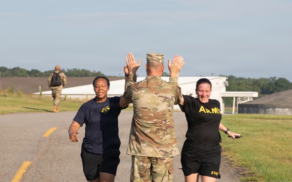 Three U.S. Army personnel are captured in a celebratory moment on a paved road at what appears to be a military installation. A soldier in camouflage uniform faces away from the camera with both hands raised, simultaneously high-fiving two female soldiers wearing Army PT (physical training) uniforms. A fourth soldier carrying a rucksack is visible in the background.