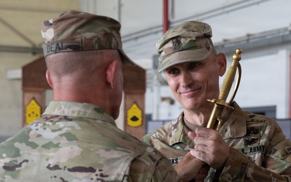 Two U.S. Army soldiers in OCP (Operational Camouflage Pattern) uniforms are engaged in what appears to be a military ceremony inside a hangar or large indoor facility. The facing soldier is holding a ceremonial saber with a gold guard, smiling, suggesting a formal presentation or award ceremony. Yellow chevron insignia boards are visible in the background, consistent with a senior NCO recognition event.