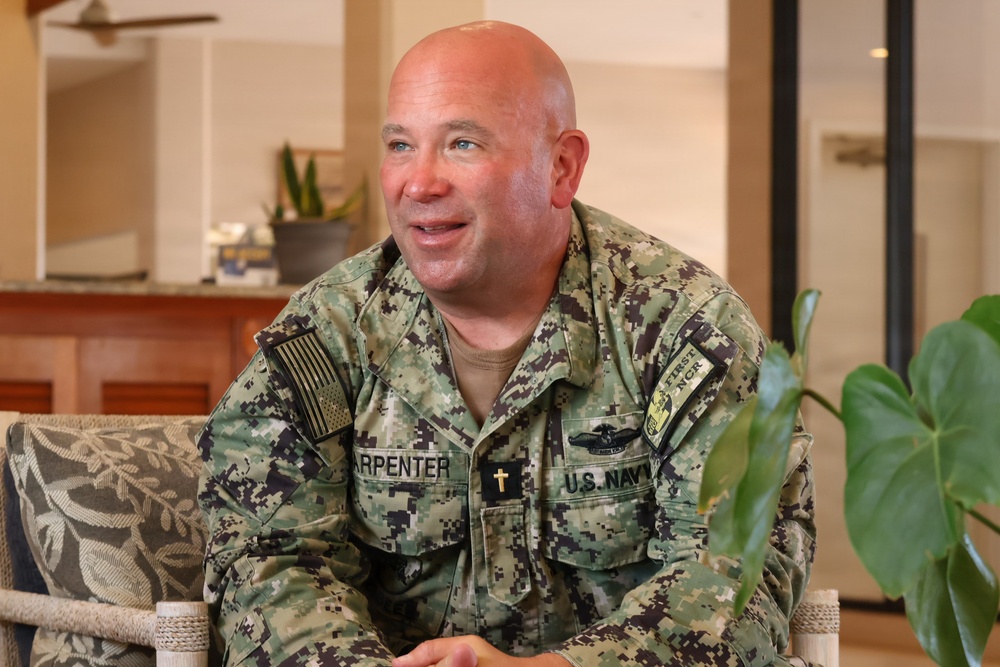 A U.S. Navy servicemember named Carpenter is seated indoors, appearing to be engaged in a conversation or interview. He is wearing the Navy Working Uniform (NWU Type III) with visible insignia including a chaplain's cross, a warfare device, and unit patches. The setting appears to be a casual indoor environment with tropical-style furnishings.