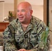A U.S. Navy servicemember named Carpenter is seated indoors, appearing to be engaged in a conversation or interview. He is wearing the Navy Working Uniform (NWU Type III) with visible insignia including a chaplain's cross, a warfare device, and unit patches. The setting appears to be a casual indoor environment with tropical-style furnishings.