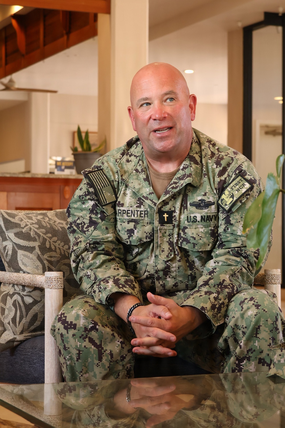 A U.S. Navy chaplain wearing NWU Type III digital camouflage uniform is seated on a couch in what appears to be a casual indoor setting. His uniform displays the name tape 'CARPENTER', a Christian cross insignia indicating chaplain status, 'U.S. NAVY' tape, a subdued American flag patch, and a 'FIRST NCR' unit patch. He appears to be engaged in conversation or an interview.