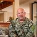 A U.S. Navy chaplain wearing NWU Type III digital camouflage uniform is seated on a couch in what appears to be a casual indoor setting. His uniform displays the name tape 'CARPENTER', a Christian cross insignia indicating chaplain status, 'U.S. NAVY' tape, a subdued American flag patch, and a 'FIRST NCR' unit patch. He appears to be engaged in conversation or an interview.