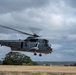 A military naval helicopter is captured in low-altitude flight or hover over an open grassy field. The aircraft is painted in a gray color scheme with 'NAVAL' markings visible on the fuselage, consistent with a Sikorsky Sea King or similar naval helicopter. The rotor blades are in motion, and debris or dust is being kicked up from the ground below.