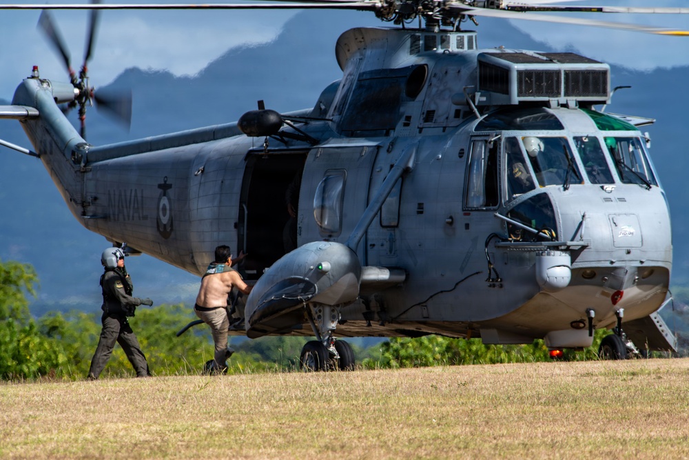 A gray military naval helicopter, marked 'NAVAL' on its fuselage, is landed on a grassy field with rotors still spinning. Two individuals are visible near the open side door — one in full tactical/flight gear and one shirtless civilian — appearing to board or disembark the aircraft. The setting appears to be a rural or semi-remote area with green hills and vegetation in the background.