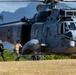 A gray military naval helicopter, marked 'NAVAL' on its fuselage, is landed on a grassy field with rotors still spinning. Two individuals are visible near the open side door — one in full tactical/flight gear and one shirtless civilian — appearing to board or disembark the aircraft. The setting appears to be a rural or semi-remote area with green hills and vegetation in the background.