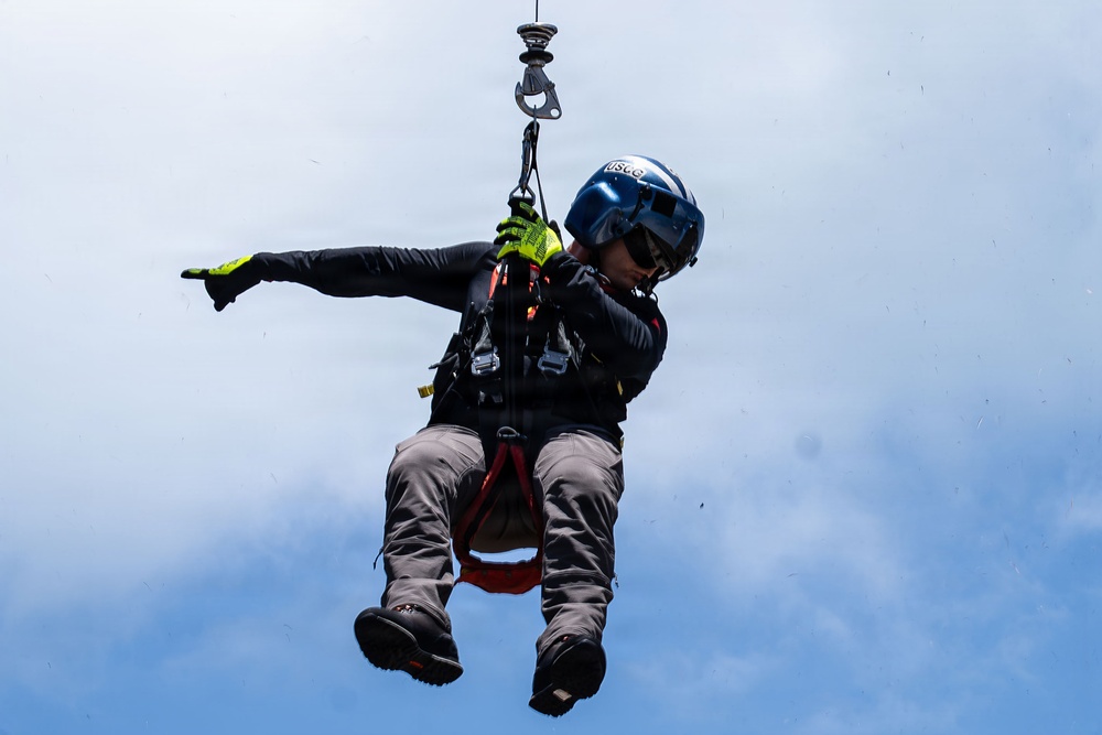 A rescue swimmer or search-and-rescue technician is suspended mid-air from a helicopter hoist cable against an overcast sky. The individual is wearing full rescue gear including a blue helmet marked 'USCG', a harness, and high-visibility gloves. This appears to be either an active rescue operation or a training exercise conducted by the United States Coast Guard.