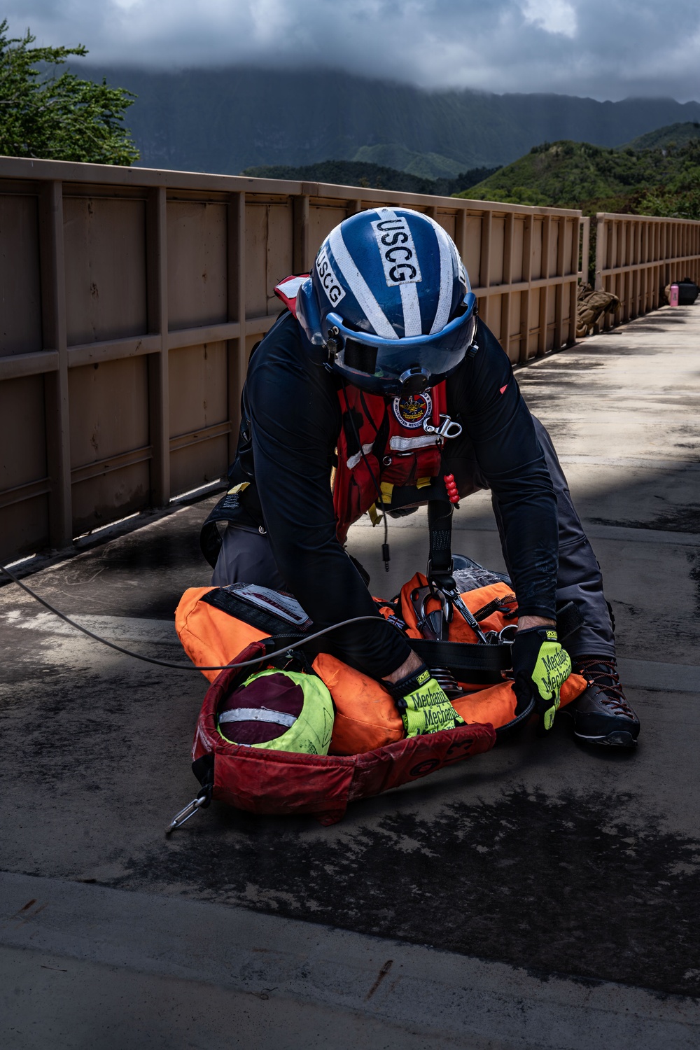 A USCG (United States Coast Guard) rescue swimmer is kneeling on what appears to be a dam or bridge surface, attending to a simulated or actual casualty secured in an orange rescue litter/basket. The rescuer is wearing full rescue gear including a blue USCG helmet, red life vest, dark dry suit, and yellow Mechanix gloves. The setting appears to be a mountainous, tropical or subtropical environment with green hills and overcast skies in the background.