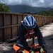 A USCG (United States Coast Guard) rescue swimmer is kneeling on what appears to be a dam or bridge surface, attending to a simulated or actual casualty secured in an orange rescue litter/basket. The rescuer is wearing full rescue gear including a blue USCG helmet, red life vest, dark dry suit, and yellow Mechanix gloves. The setting appears to be a mountainous, tropical or subtropical environment with green hills and overcast skies in the background.