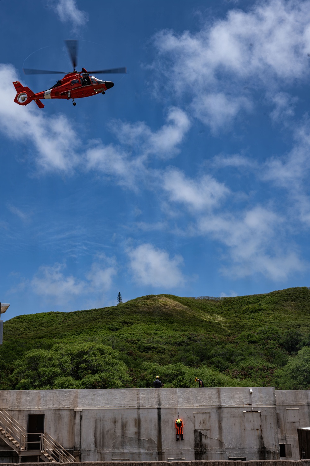 A red U.S. Coast Guard helicopter hovers above a large concrete structure, likely a dam or flood control facility, while personnel conduct what appears to be a rescue or training exercise. One person in an orange safety harness is rappelling or suspended on the vertical concrete wall, while two other individuals stand on top of the structure observing. The scene is set against a lush green hillside under a partly cloudy blue sky.