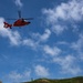 A red U.S. Coast Guard helicopter hovers above a large concrete structure, likely a dam or flood control facility, while personnel conduct what appears to be a rescue or training exercise. One person in an orange safety harness is rappelling or suspended on the vertical concrete wall, while two other individuals stand on top of the structure observing. The scene is set against a lush green hillside under a partly cloudy blue sky.