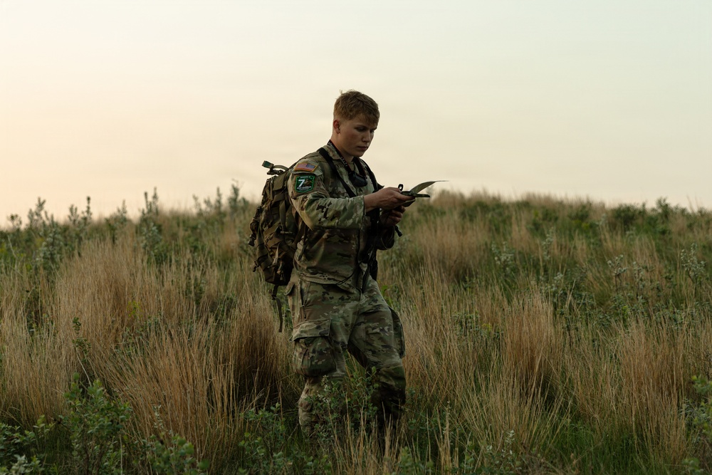 A young soldier in multicam/OCP camouflage uniform stands in an open grassy field during low-light conditions, appearing to consult a map or document. He is equipped with a tactical backpack and chest rig, and wears unit patches on his left shoulder. The setting suggests a land navigation or field training exercise.