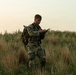 A young soldier in multicam/OCP camouflage uniform stands in an open grassy field during low-light conditions, appearing to consult a map or document. He is equipped with a tactical backpack and chest rig, and wears unit patches on his left shoulder. The setting suggests a land navigation or field training exercise.