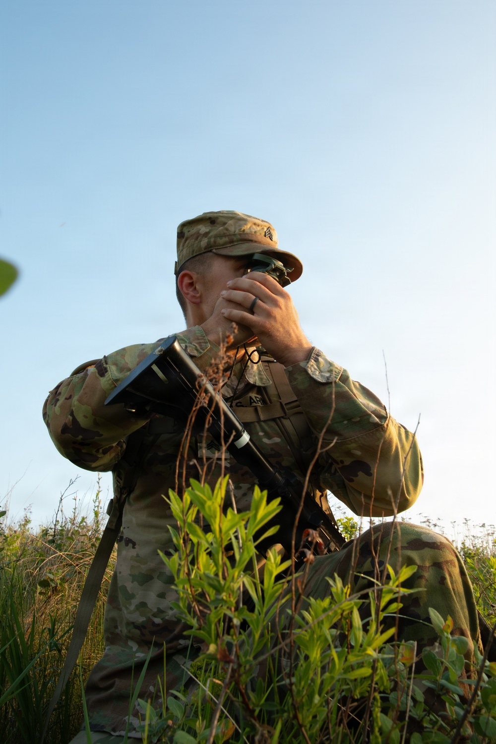 A U.S. Army Sergeant in Operational Camouflage Pattern (OCP) uniform is positioned in a field using binoculars or a monocular device for observation. The soldier is carrying a rifle slung across their body and is partially concealed by low vegetation. The image is taken from a low angle during golden hour lighting conditions.