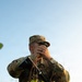 A U.S. Army Sergeant in Operational Camouflage Pattern (OCP) uniform is positioned in a field using binoculars or a monocular device for observation. The soldier is carrying a rifle slung across their body and is partially concealed by low vegetation. The image is taken from a low angle during golden hour lighting conditions.