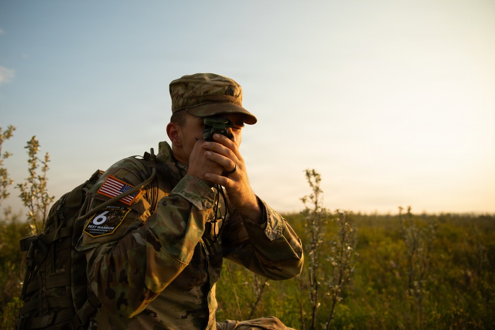 A U.S. Army soldier in MultiCam (OCP) camouflage uniform is using a handheld optical device, likely a lensatic compass or rangefinder, in an outdoor field setting during golden hour lighting. The soldier is wearing a patrol cap with sergeant rank insignia and a rucksack bearing a U.S. flag patch and a 'Region VI Best Warrior Competition' patch. The scene suggests a land navigation or reconnaissance exercise.