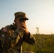 A U.S. Army soldier in MultiCam (OCP) camouflage uniform is using a handheld optical device, likely a lensatic compass or rangefinder, in an outdoor field setting during golden hour lighting. The soldier is wearing a patrol cap with sergeant rank insignia and a rucksack bearing a U.S. flag patch and a 'Region VI Best Warrior Competition' patch. The scene suggests a land navigation or reconnaissance exercise.