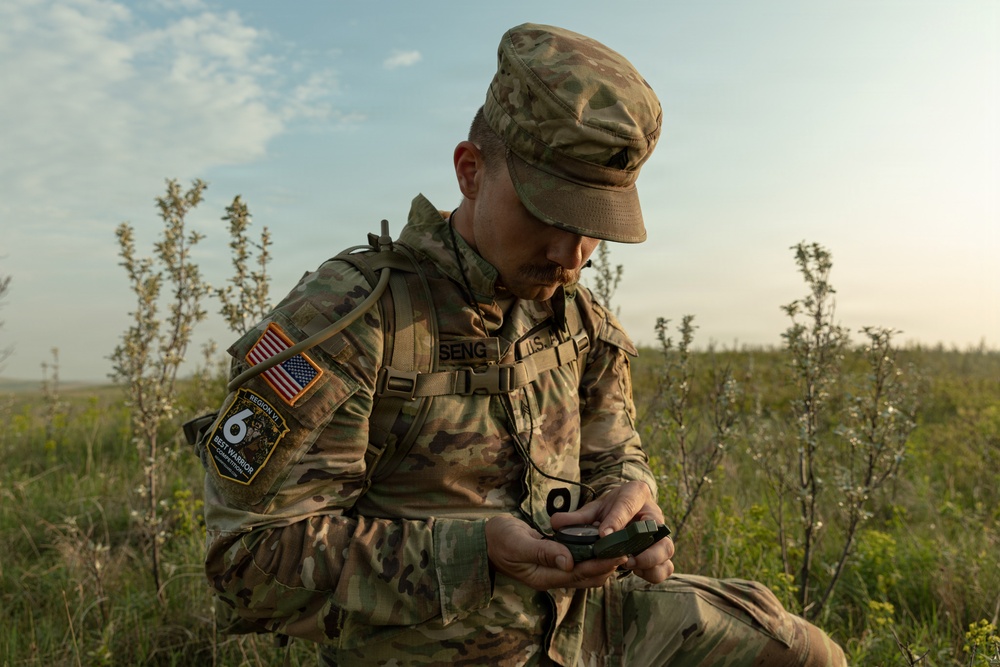 A U.S. Army soldier in full Operational Camouflage Pattern (OCP) uniform is seated in an open field, examining a military compass. The soldier is wearing a patrol cap and tactical backpack, with visible patches including an American flag and a 'Region VI Best Warrior Competition' patch. The setting appears to be a training or field exercise environment during daylight hours.