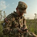 A U.S. Army soldier in full Operational Camouflage Pattern (OCP) uniform is seated in an open field, examining a military compass. The soldier is wearing a patrol cap and tactical backpack, with visible patches including an American flag and a 'Region VI Best Warrior Competition' patch. The setting appears to be a training or field exercise environment during daylight hours.