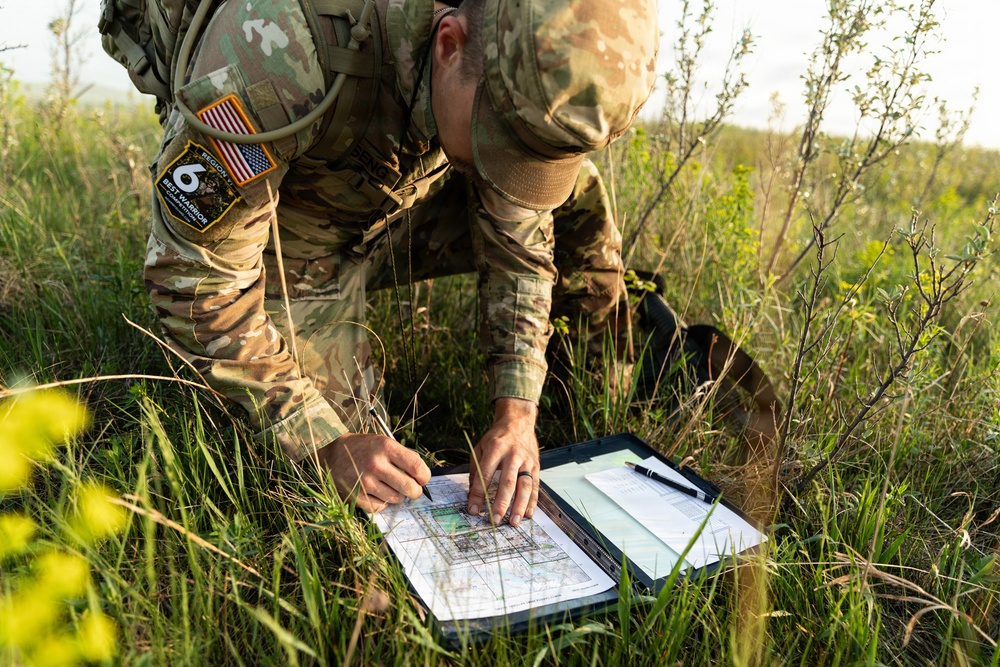 A U.S. Army soldier in MultiCam/OCP camouflage uniform is kneeling in a grassy field, studying a topographic map on a clipboard. The soldier is wearing a tactical vest with visible patches including a U.S. flag patch and a 'Region 6 Best Warrior' patch. This appears to be a land navigation exercise or field training activity.