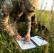 A U.S. Army soldier in MultiCam/OCP camouflage uniform is kneeling in a grassy field, studying a topographic map on a clipboard. The soldier is wearing a tactical vest with visible patches including a U.S. flag patch and a 'Region 6 Best Warrior' patch. This appears to be a land navigation exercise or field training activity.
