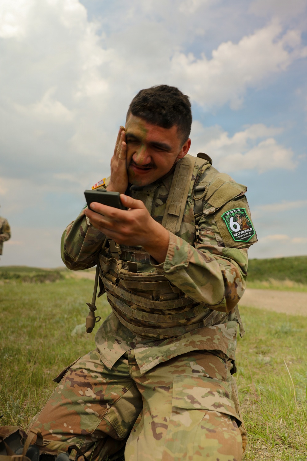 A soldier in U.S. Army OCP (Operational Camouflage Pattern) uniform is kneeling outdoors and applying camouflage face paint while using a smartphone as a mirror. He is wearing a plate carrier vest and has a patch on his left sleeve reading 'Region VI Best Warrior Competition.' The setting appears to be an open field or training area under a partly cloudy sky.