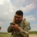 A soldier in U.S. Army OCP (Operational Camouflage Pattern) uniform is kneeling outdoors and applying camouflage face paint while using a smartphone as a mirror. He is wearing a plate carrier vest and has a patch on his left sleeve reading 'Region VI Best Warrior Competition.' The setting appears to be an open field or training area under a partly cloudy sky.