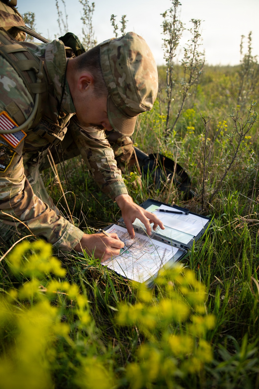 A U.S. Army soldier in OCP (Operational Camouflage Pattern) uniform is crouched in an open grassy field conducting land navigation, studying a topographic map on a field clipboard. The soldier wears a U.S. flag patch on the left shoulder and carries a tactical backpack. The setting appears to be a training exercise in a natural outdoor environment during daylight hours.