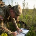 A U.S. Army soldier in OCP (Operational Camouflage Pattern) uniform is crouched in an open grassy field conducting land navigation, studying a topographic map on a field clipboard. The soldier wears a U.S. flag patch on the left shoulder and carries a tactical backpack. The setting appears to be a training exercise in a natural outdoor environment during daylight hours.