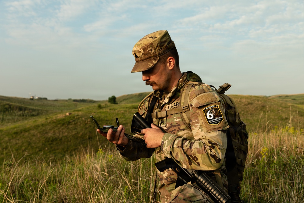 A U.S. Army soldier in MultiCam (OCP) camouflage uniform is standing in an open grassy field while reading a compass. He is wearing a tactical vest and carrying a rifle, with a visible patch identifying him as a participant in the 'Region VI Best Warrior Competition.' The setting appears to be a rural, hilly landscape under a partly cloudy sky.