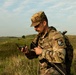 A U.S. Army soldier in MultiCam (OCP) camouflage uniform is standing in an open grassy field while reading a compass. He is wearing a tactical vest and carrying a rifle, with a visible patch identifying him as a participant in the 'Region VI Best Warrior Competition.' The setting appears to be a rural, hilly landscape under a partly cloudy sky.