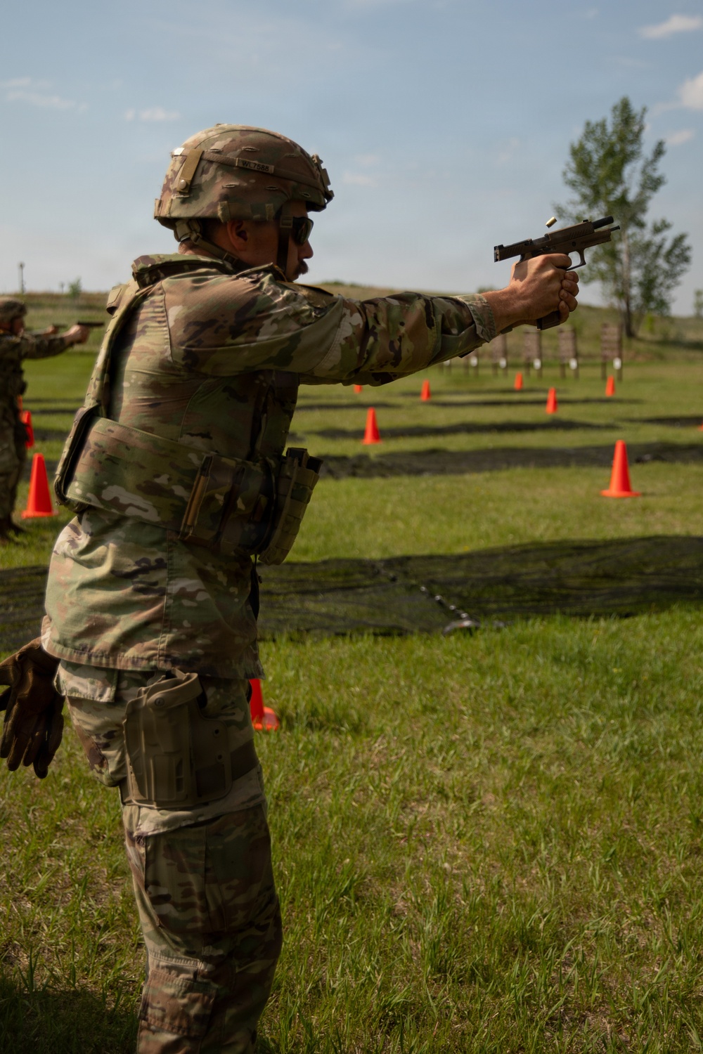 A military service member in full OCP (Operational Camouflage Pattern) uniform and body armor is firing a pistol on an outdoor shooting range. A spent brass casing is visibly ejecting from the firearm. Additional personnel are visible in the background conducting similar firing exercises.