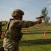A military service member in full OCP (Operational Camouflage Pattern) uniform and body armor is firing a pistol on an outdoor shooting range. A spent brass casing is visibly ejecting from the firearm. Additional personnel are visible in the background conducting similar firing exercises.