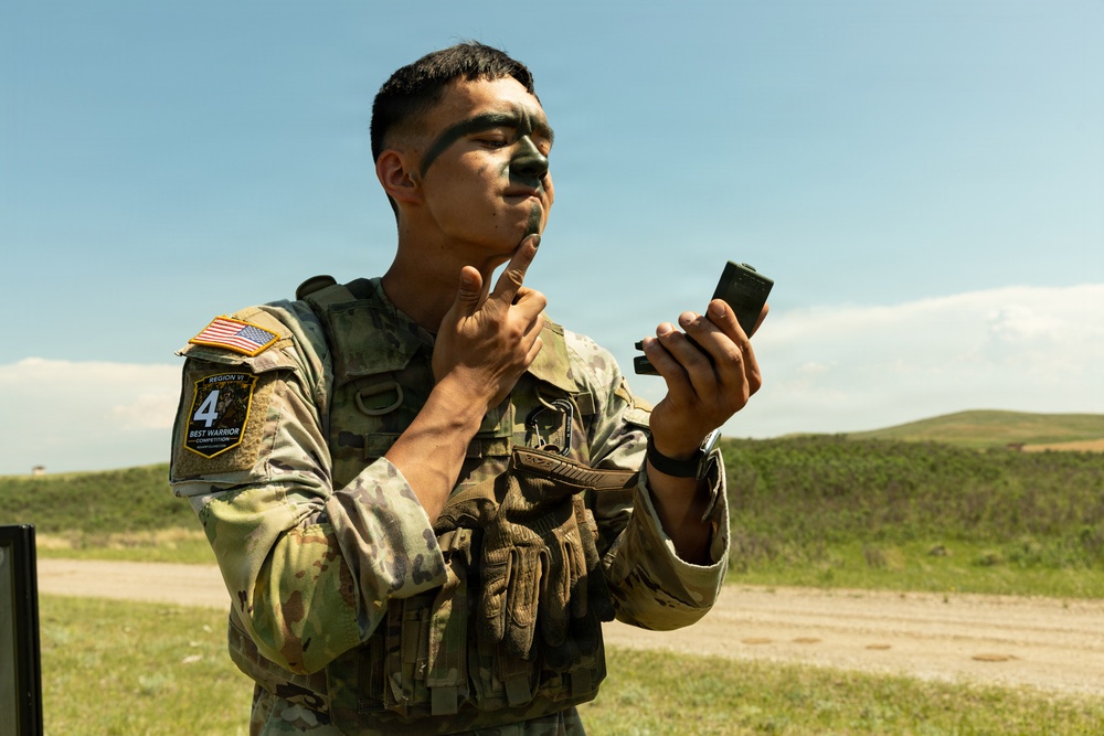 A U.S. Army soldier in OCP (Operational Camouflage Pattern) uniform is applying camouflage face paint while looking into a small compact mirror. He is wearing a tactical vest with a visible patch reading 'Region VI Best Warrior Competition' with the number 4, along with a U.S. flag patch. The setting appears to be an outdoor training or competition area with rolling green hills and a dirt road in the background.