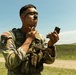A U.S. Army soldier in OCP (Operational Camouflage Pattern) uniform is applying camouflage face paint while looking into a small compact mirror. He is wearing a tactical vest with a visible patch reading 'Region VI Best Warrior Competition' with the number 4, along with a U.S. flag patch. The setting appears to be an outdoor training or competition area with rolling green hills and a dirt road in the background.