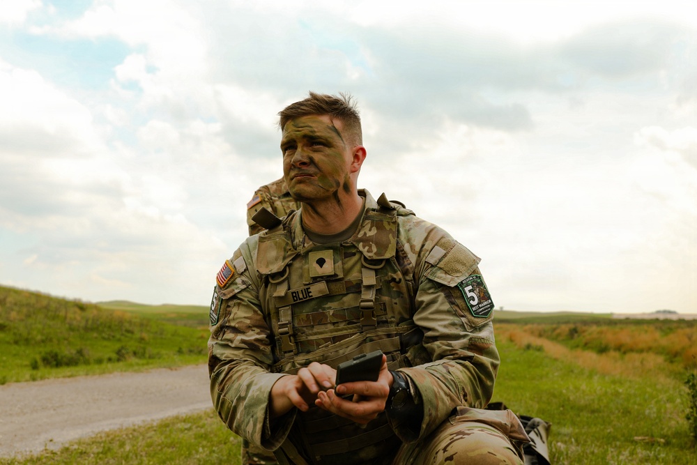 A U.S. Army soldier in MultiCam (OCP) camouflage uniform is seated outdoors in a grassy field area, wearing tactical gear and face camouflage paint. The soldier, identified by a name tape reading 'BLUE,' is holding a small electronic device and appears to be resting or taking a break during a field exercise or competition. A patch on the right shoulder indicates participation in what appears to be a 'Best Warrior' competition.