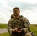 A U.S. Army soldier in MultiCam (OCP) camouflage uniform is seated outdoors in a grassy field area, wearing tactical gear and face camouflage paint. The soldier, identified by a name tape reading 'BLUE,' is holding a small electronic device and appears to be resting or taking a break during a field exercise or competition. A patch on the right shoulder indicates participation in what appears to be a 'Best Warrior' competition.