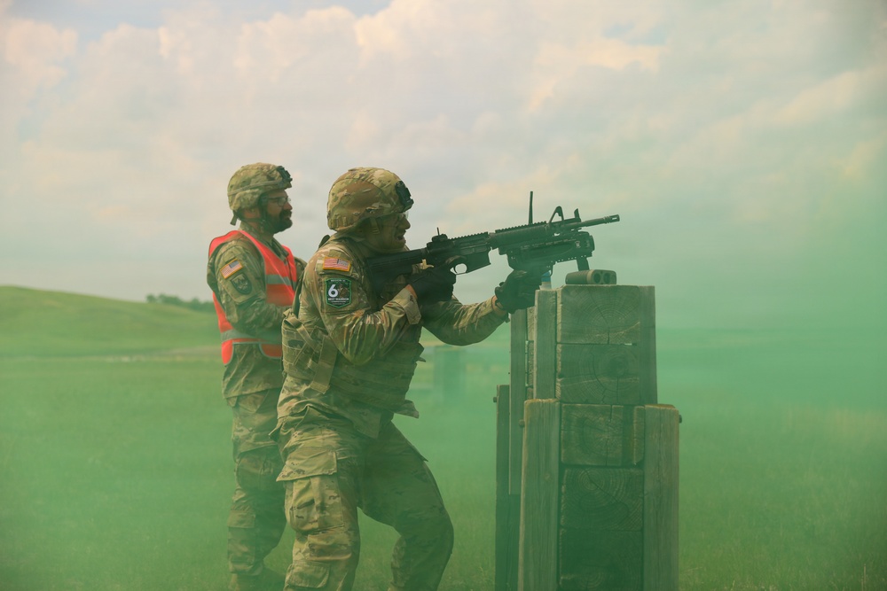 Two soldiers in U.S. Army camouflage uniforms are engaged in what appears to be a military training exercise on an open field. One soldier is firing or aiming a rifle (appears to be an M4/M16-style weapon) from behind a concrete barrier, while a second soldier wearing a high-visibility orange safety vest observes or supervises. Green smoke from a smoke grenade fills the surrounding area, indicating a tactical training scenario.